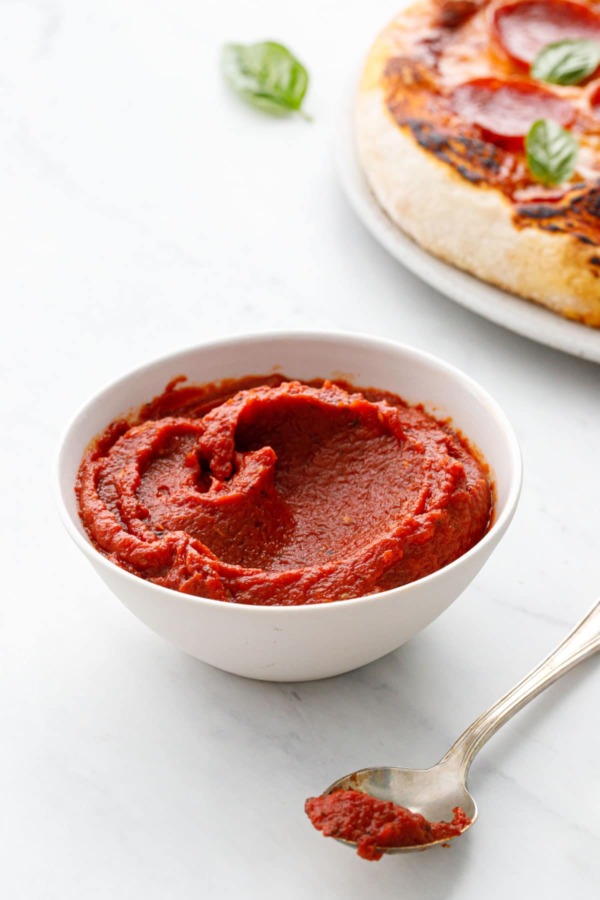 White bowl with homemade pizza sauce and spoon, homemade pizza on a plate in the background