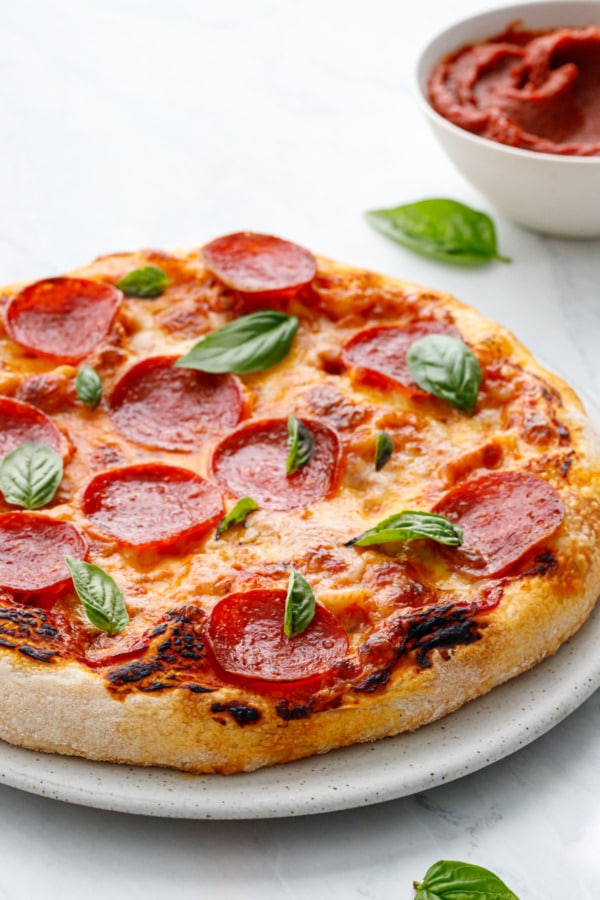 Freshly baked pepperoni and cheese pizza with fresh basil leaves with bowl of homemade pizza sauce in background