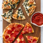 Overhead wooden cutting board with two sliced pizzas (pepperoni and pizza bianca), with a bowl of sauce
