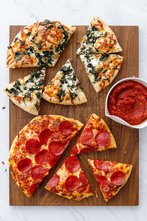 Overhead wooden cutting board with two sliced pizzas (pepperoni and pizza bianca), with a bowl of sauce