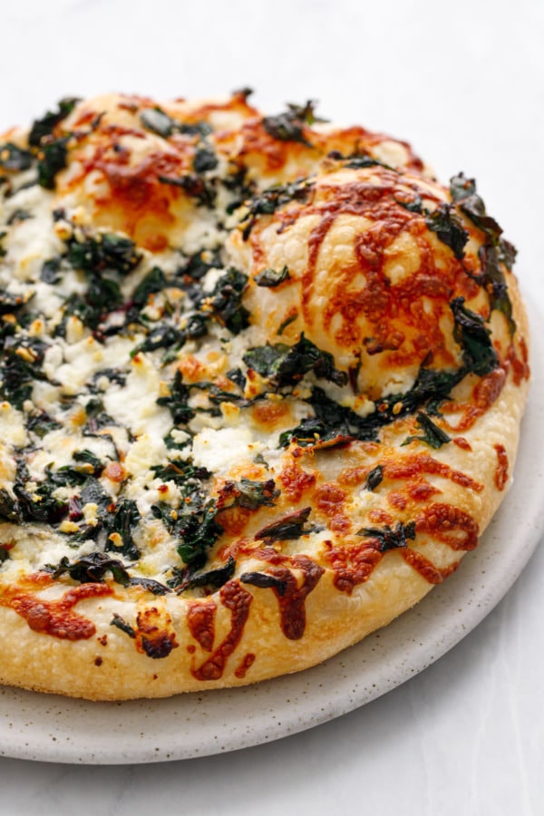 Closeup of a baked pizza topped with greens and goat cheese, with browned shreds of cheese and a big bubble
