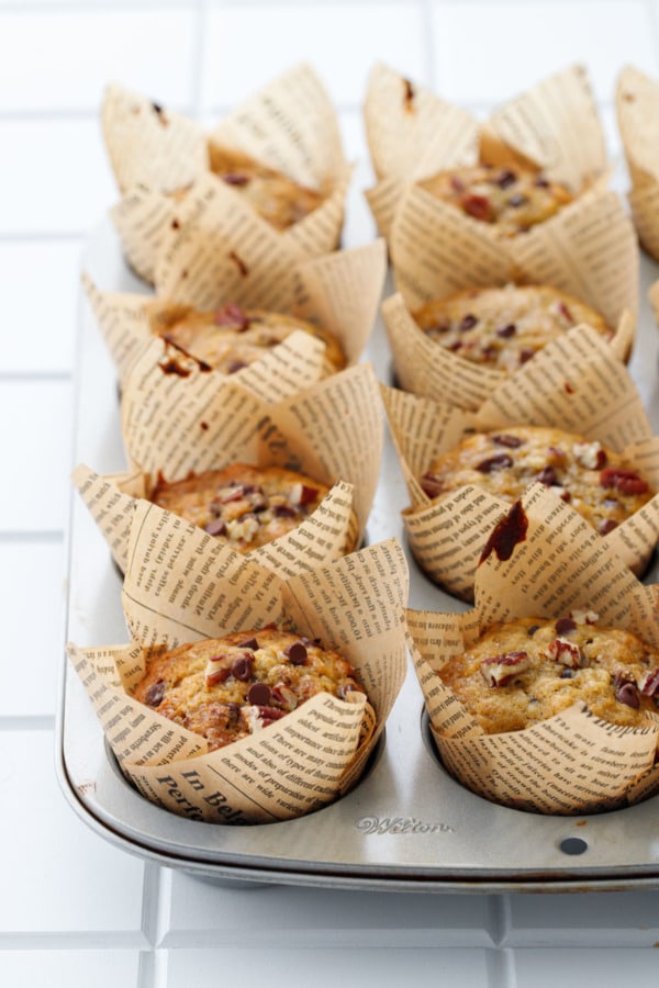 Silver muffin tin with newsprint tulip wrappers, baked banana bread muffins on a white background