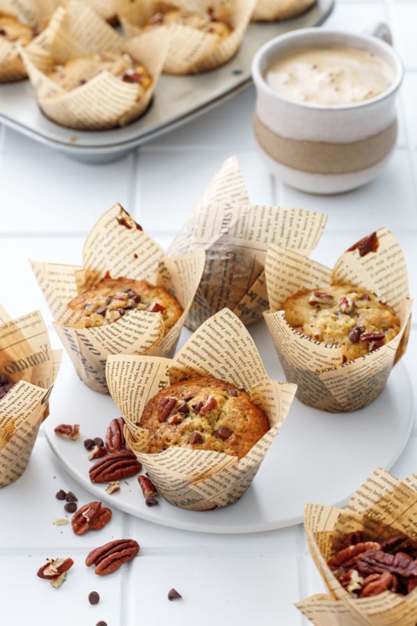 White tile background with banana bread muffins in tulip papers, cup of coffee and muffin tin in the background