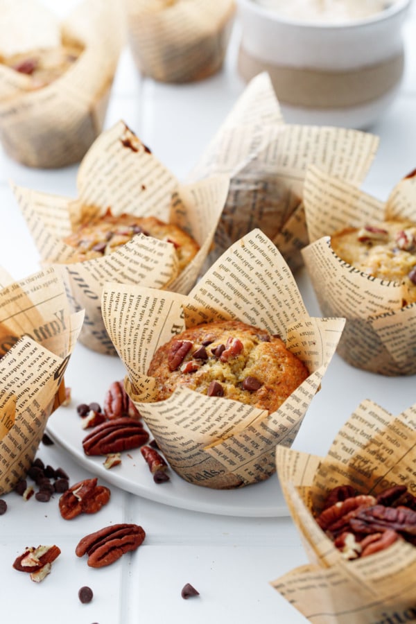 White plate with banana bread muffins, loose pecans and chocolate chips and a cup of coffee in the backgorund