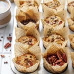 Muffin tin with banana bread muffins, one cup filled with raw pecans and one with chocolate chips, and cup of coffee