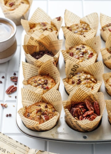 Muffin tin with banana bread muffins, one cup filled with raw pecans and one with chocolate chips, and cup of coffee