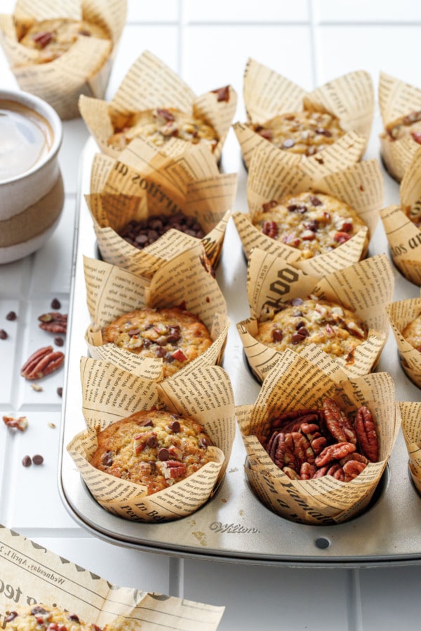 Muffin tin with banana bread muffins, one cup filled with raw pecans and one with chocolate chips, and cup of coffee