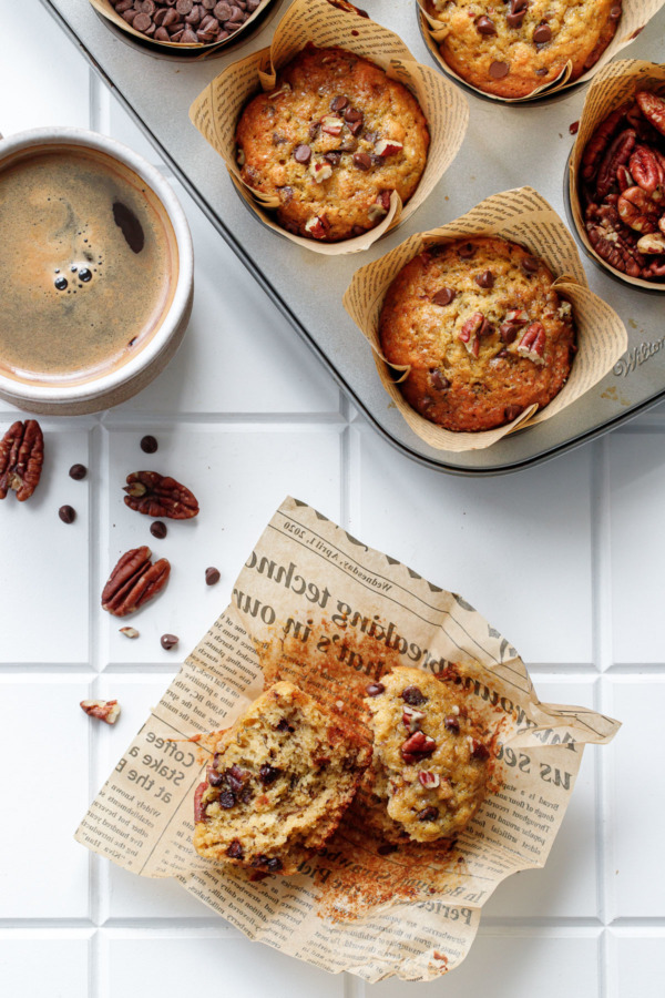 Overhead, white tile background with peeled muffin split in half to show texture, muffin tin and cup of coffee