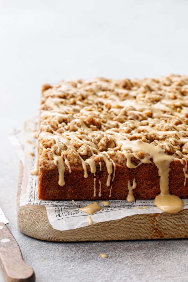 Wood board with Espresso Crumb Coffee Cake, drizzled with coffee glaze dripping down the sides and newsprint parchment paper