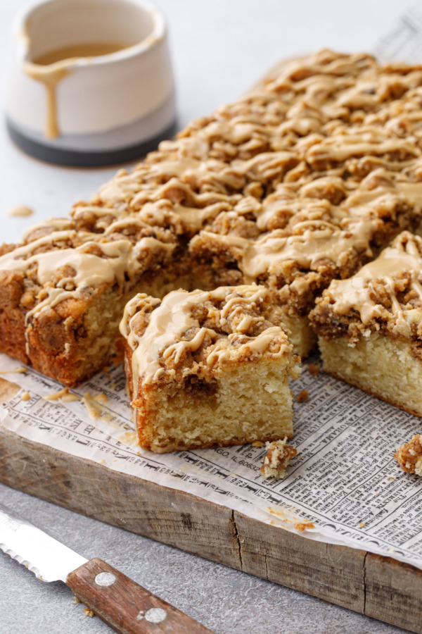 Espresso Crumb Coffee Cake cut into squares, on a wood board with newsprint parchment paper, cup of glaze in the background