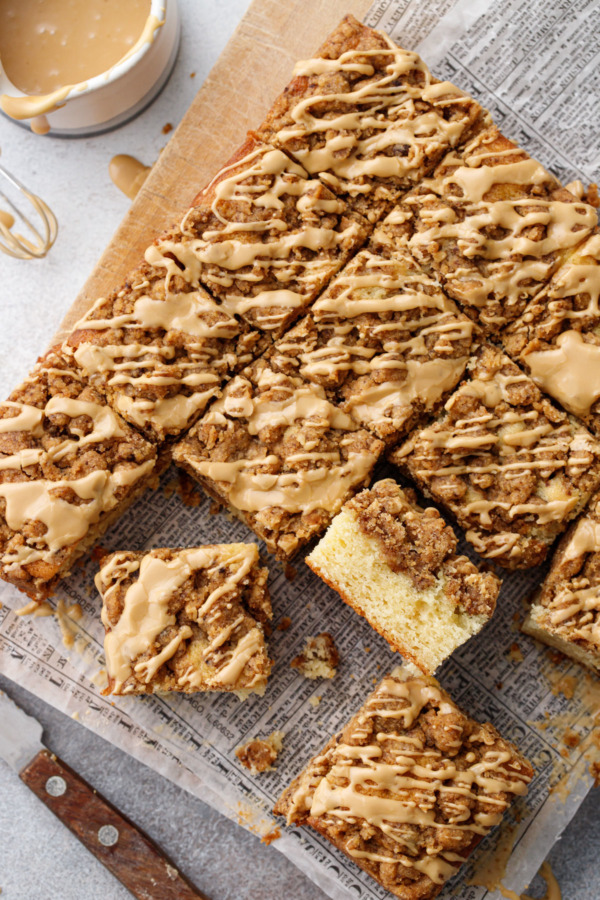 Overhead, offset grid of coffee cake squares, on a wood board with creamer filled with glaze and wooden-handled knife