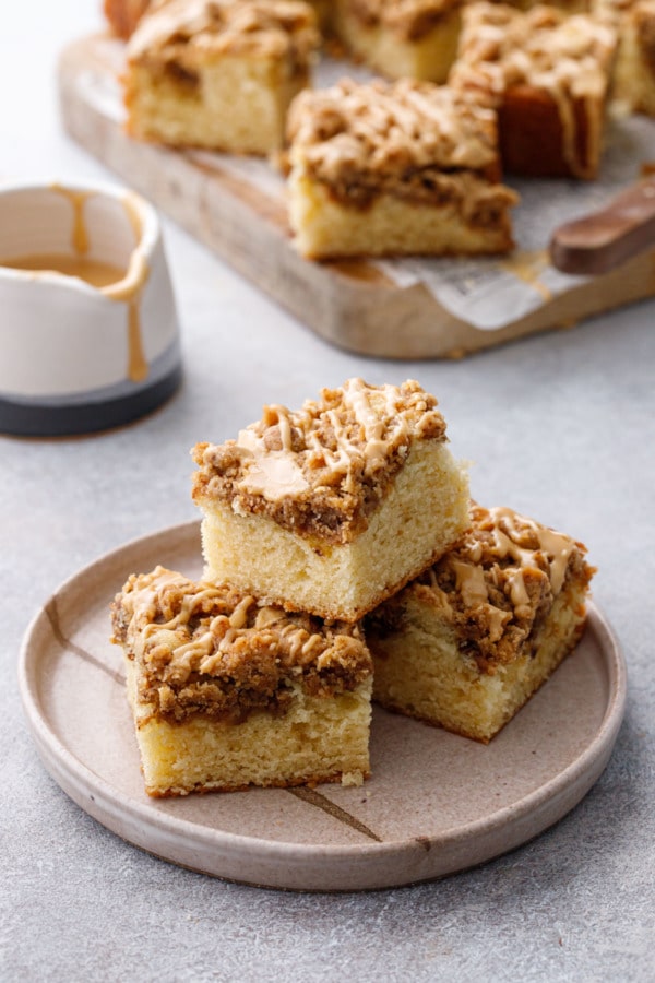 Ceramic plate with pyramid stack of coffee cake squares, wooden cutting board with more slices and cup of glaze in the background