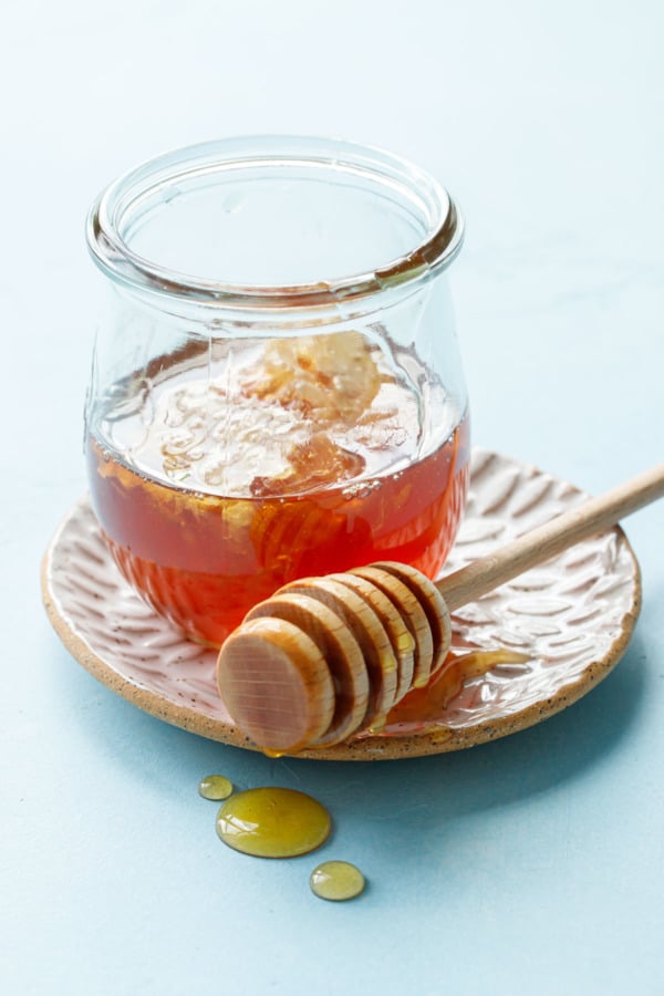 Glass jar of honey with wooden drizzler on a homemade ceramic plate, blue background