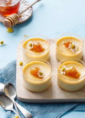 Four glass ramekins with honey pots de creme on a wood board, light blue background with a jar of honey and spoons