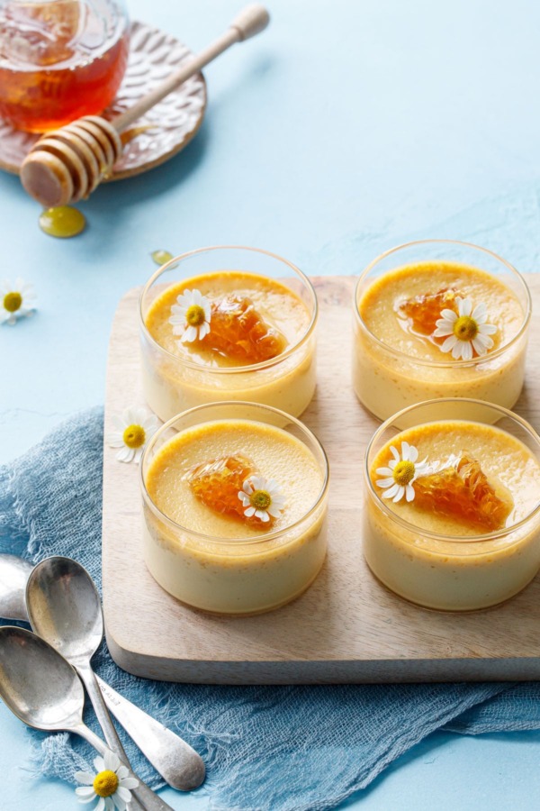 Four glass ramekins with honey pots de creme on a wood board, light blue background with a jar of honey and spoons