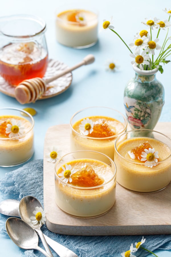 Honey pots de creme in glass ramekins on a wood board, small vase with chamomile flowers and a jar of honey in the background