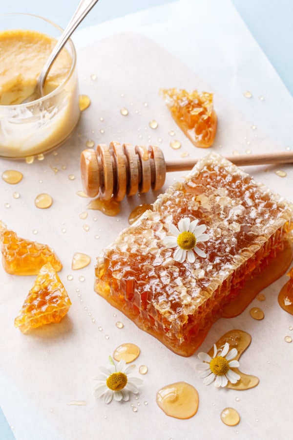 Parchment surface with piece of honeycomb, chamomile flowers, and a wooden honey drizzler