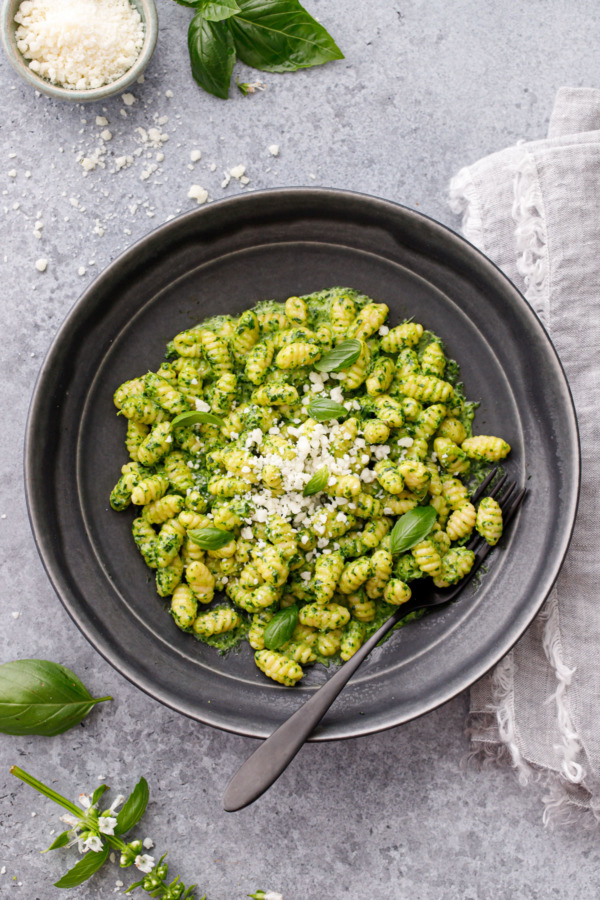 Overhead, shallow black pasta bowl with homemade gnochetti sardi in pesto sauce, with linen, grated cheese and basil leaves