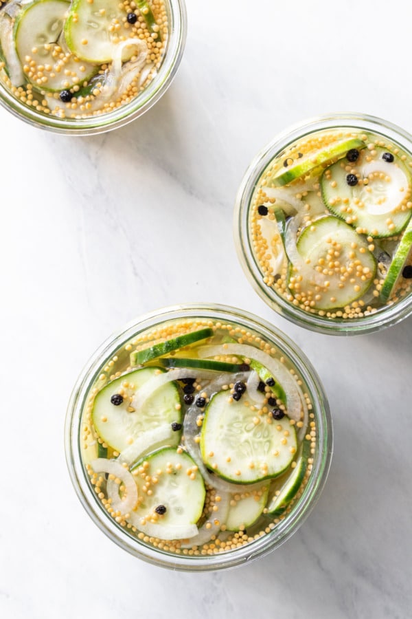 Overhead, three jars filled with Bread & Butter Refrigerator Pickles, showing the whole mustard seeds, pepper corns, and onions with the cucumber slices.