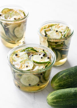 Three uncovered jars of Bread & Butter Refrigerator Pickles in brine, with two kirby cucumbers on the side