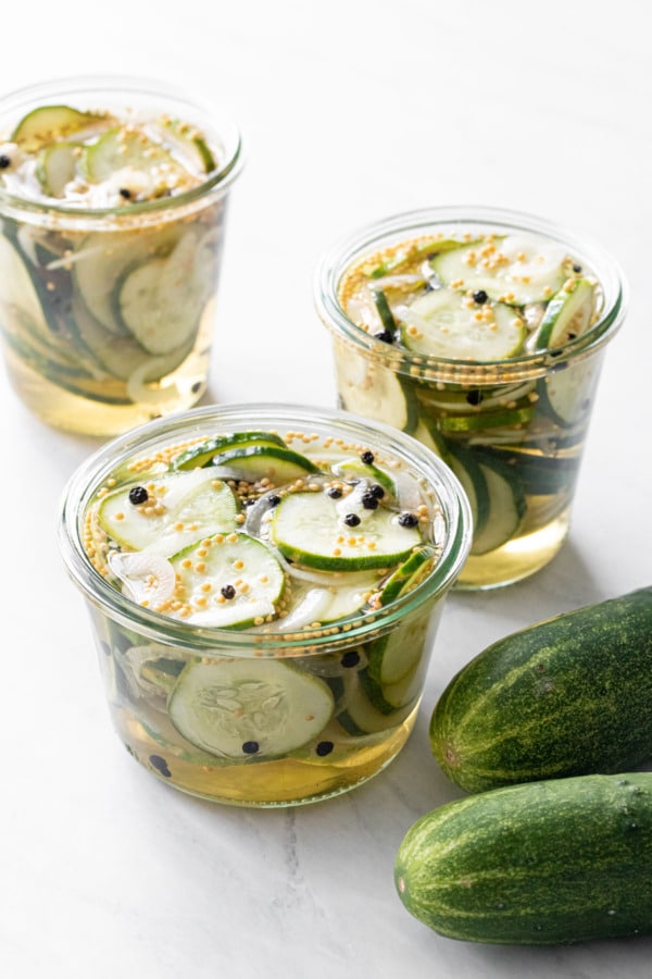 Three uncovered jars of Bread & Butter Refrigerator Pickles in brine, with two kirby cucumbers on the side