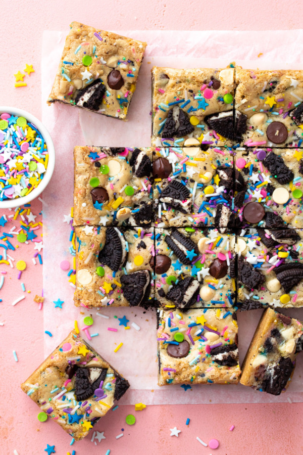 Overhead, squares of sprinkle cookie party blondies on a pink background, with bowl of sprinkles on the side