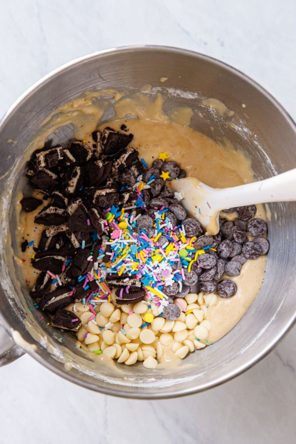 Overhead, bowl of raw blondie batter with piles of cookies, sprinkles, white and dark chocolate chips before mixing