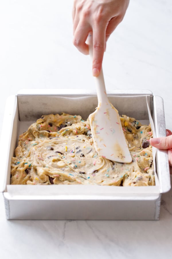 Using a spatula to spread the thick blondie batter into a parchment-lined baking pan