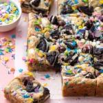Squares of sprinkle cookie party blondies on a pink background, with bowl of sprinkles on the side