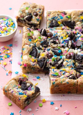 Squares of sprinkle cookie party blondies on a pink background, with bowl of sprinkles on the side