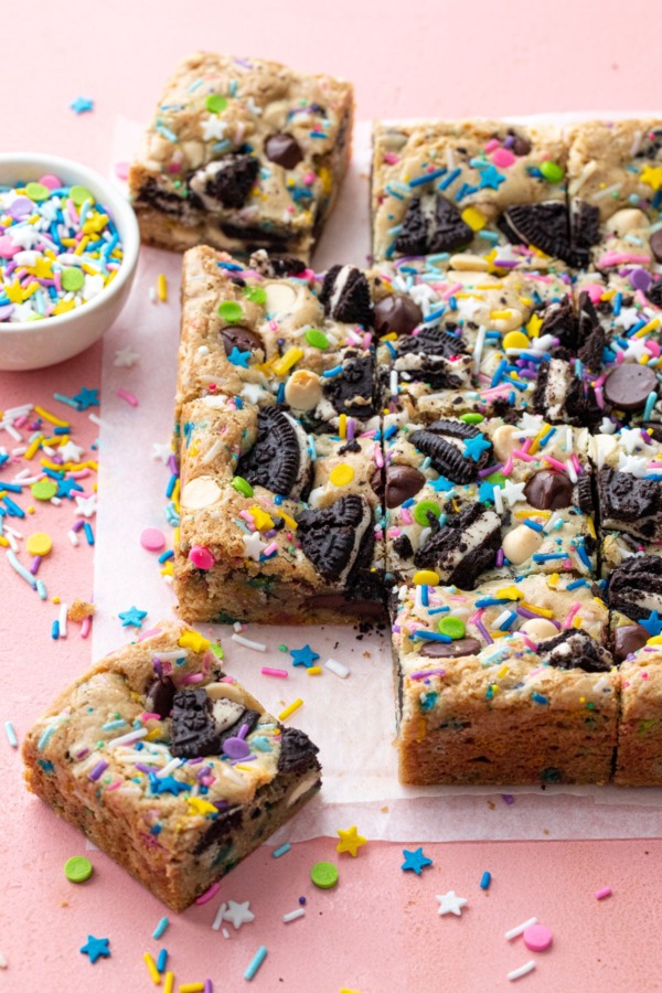 Squares of sprinkle cookie party blondies on a pink background, with bowl of sprinkles on the side