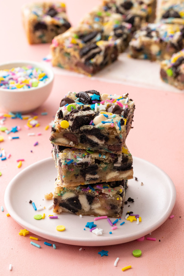 Stack of three cut squares of Sprinkle Party Blondies, on a pink plate with sprinkles in the background