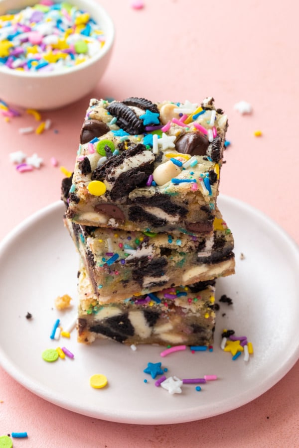 Stack of three sprinkle party blondies with sandwich cookies, on a pink plate and pink background