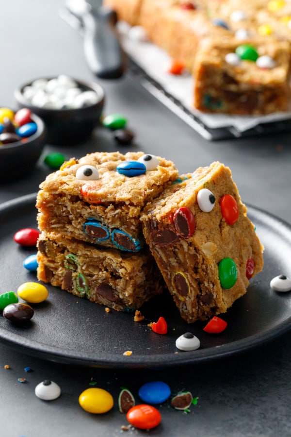 Stack of three Monster Cookie Bars on a black plate, topped with M&Ms and candy eyeballs for halloween