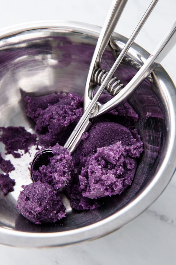 Silver mixing bowl with bright purple ube amaretti cookie dough and a metal cookie scoop