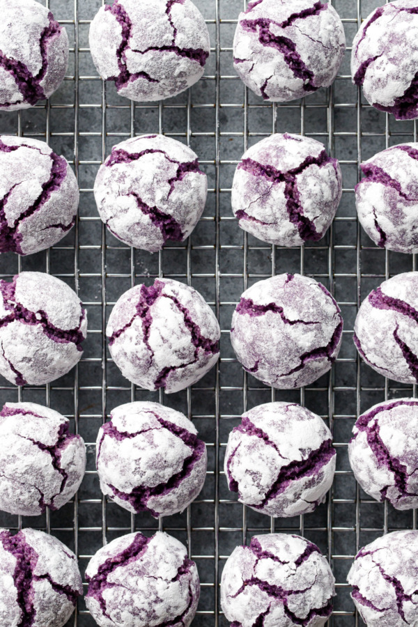 Overhead, wire rack with even rows of ube ameretti cookies, with purple cracks showing through a powdered sugar coating