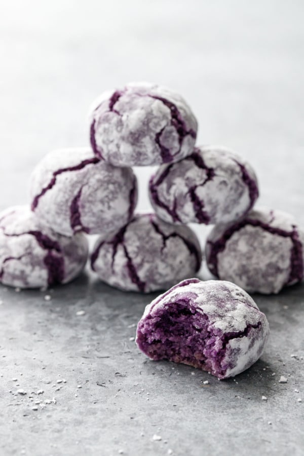 Stack of purple ube amaretti crinkle cookies on gray background, bite out of one cookie in the foreground
