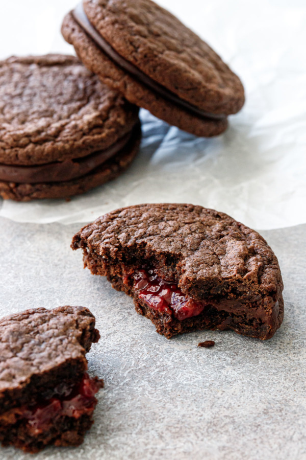 Three Chocolate Raspberry Sandwich Cookies on crinkled parchment, one cookie broken into pieces with raspberry jam oozing out of the middle.