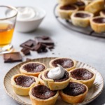 Stack of Mini Bourbon Fudge Pies on a plate, one topped with a swirl of whipped cream, shot of bourbon and bowl of whipped cream in the background