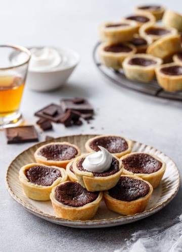 Stack of Mini Bourbon Fudge Pies on a plate, one topped with a swirl of whipped cream, shot of bourbon and bowl of whipped cream in the background