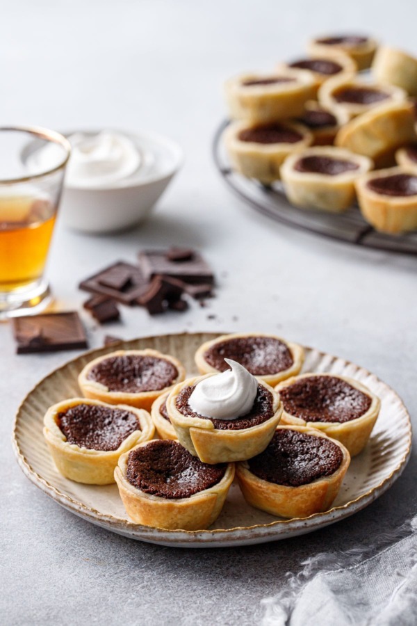 Stack of Mini Bourbon Fudge Pies on a plate, one topped with a swirl of whipped cream, shot of bourbon and bowl of whipped cream in the background