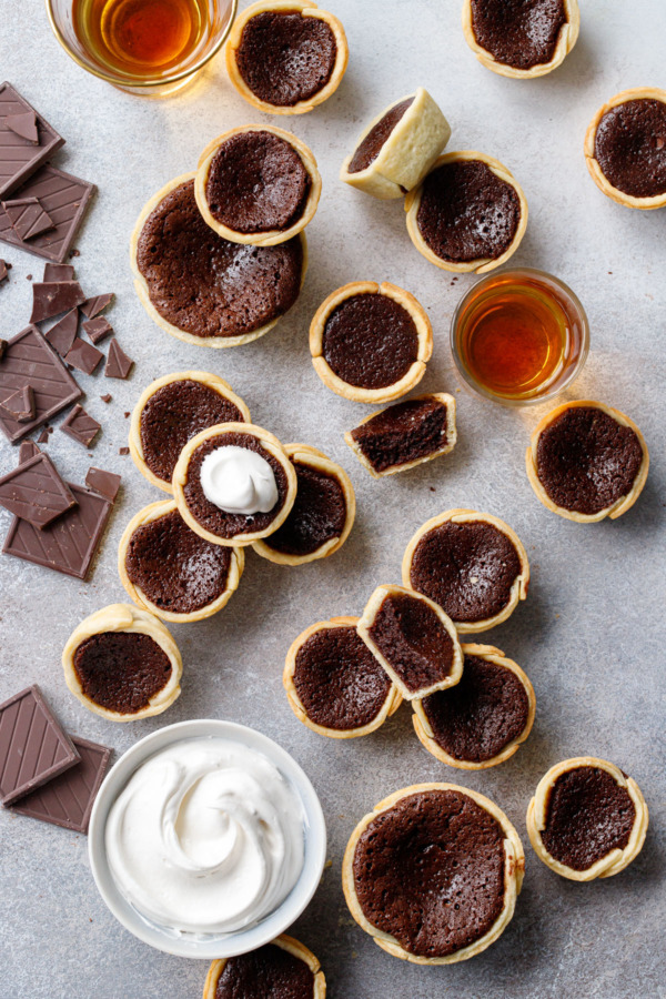 Overhead, messy arrangement of Mini Bourbon Fudge Pies on a gray background, with broken chocolate, shot of bourbon, and bowl of whipped cream