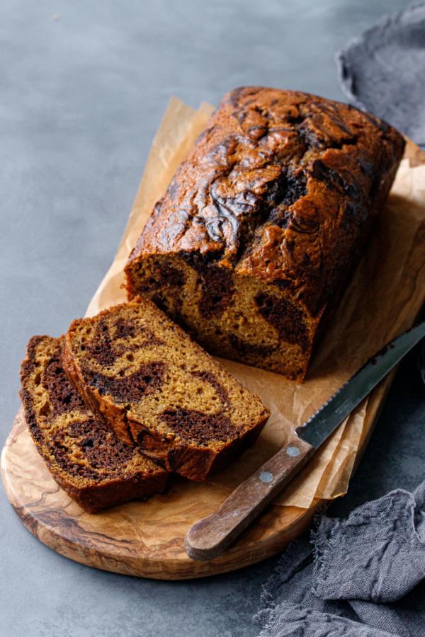 Sliced loaf of Pumpkin Chocolate Swirl Bread on an olivewood bread board with knife, dark gray background