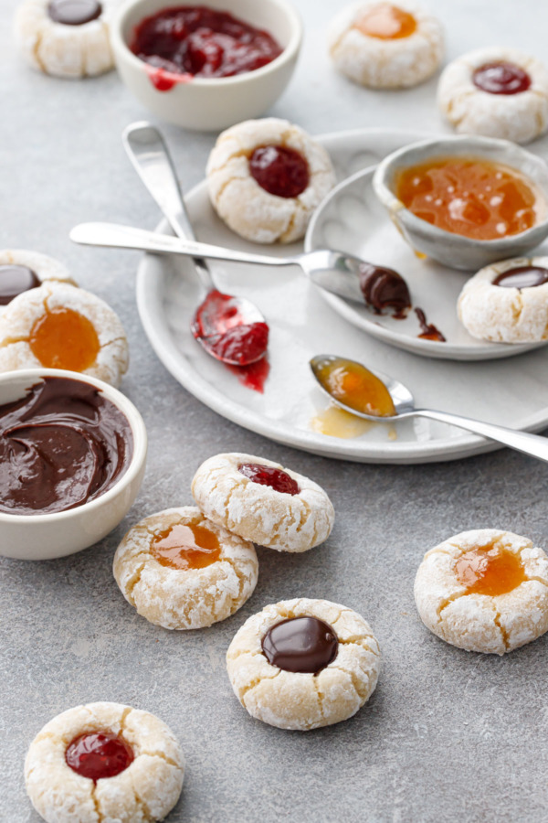 Amaretto Amaretti Thumbprint cookies on a gray background, with bowls of the various filling options messily scattered around