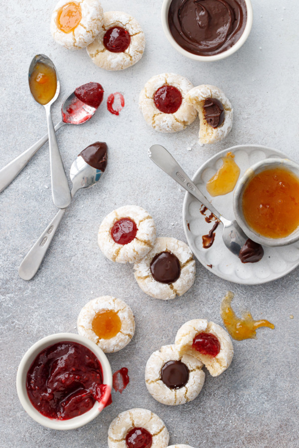 Overhead, Messy Amaretto Amaretti Thumbprint cookies on a gray background, with bowls of the various fillings scattered around