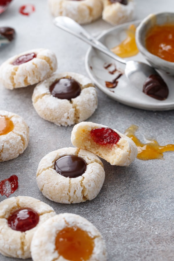 Messy arrangement of Amaretto Amaretti Thumbprint cookies, smears of jam and spoons on the side. One cookie with a bite out of it to show texture.