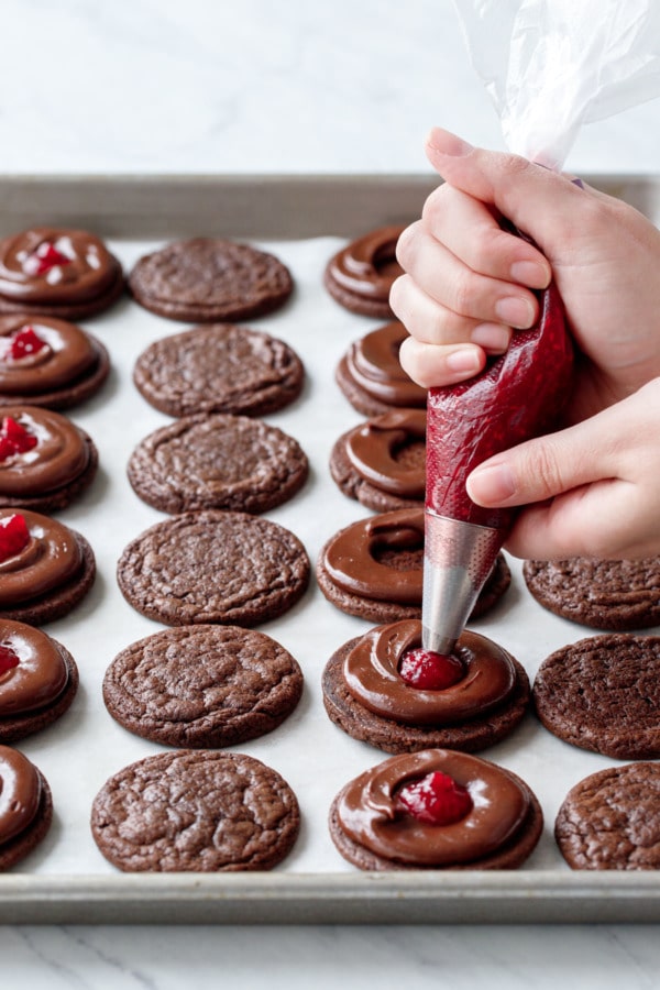 Pan with rows of chocolate brownie cookies, piping raspberry jam into the center of a ring of chocolate ganache