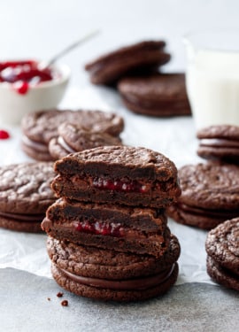 Chocolate Raspberry Sandwich Cookies, stack of cookies with one cut in half to show the fillings, with glass of milk and raspberry jam in the background.