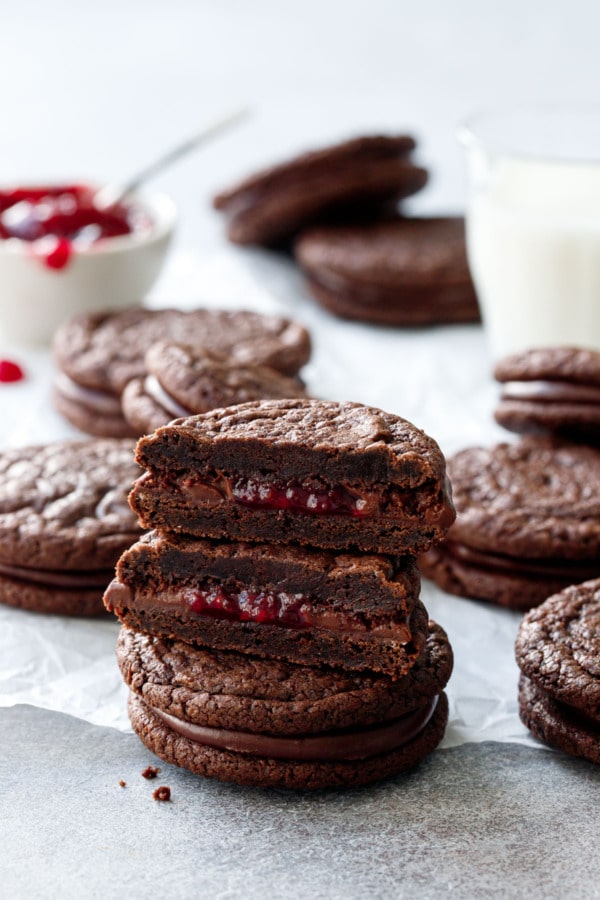 Chocolate Raspberry Sandwich Cookies, stack of cookies with one cut in half to show the fillings, with glass of milk and raspberry jam in the background.