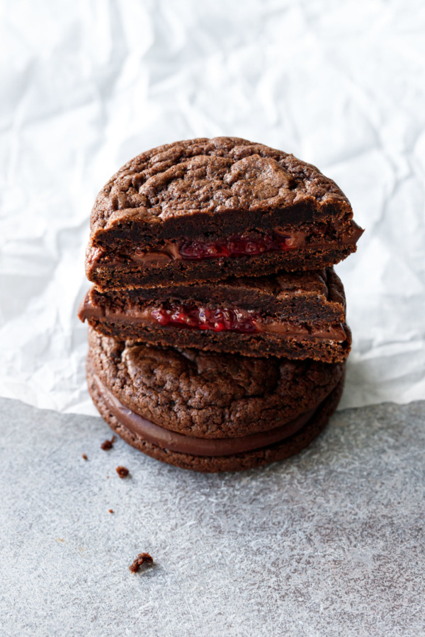 Stack of Chocolate Raspberry Sandwich Cookies on a crinkled parchment and gray background, one cookie cut in half to show the core of raspberry jam.